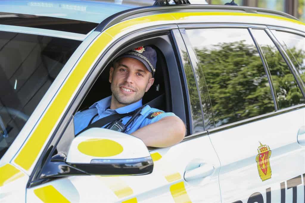 Bildet viser en smilende, ung norsk politimann sittende i tjenestebil med vinduet åpent.
