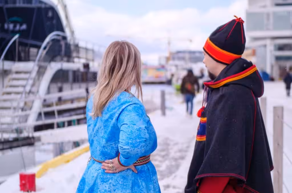 En samisk mann og kvinne i tradisjonelle klesplagg, står ved en havn og ser folk gå langs havnen.