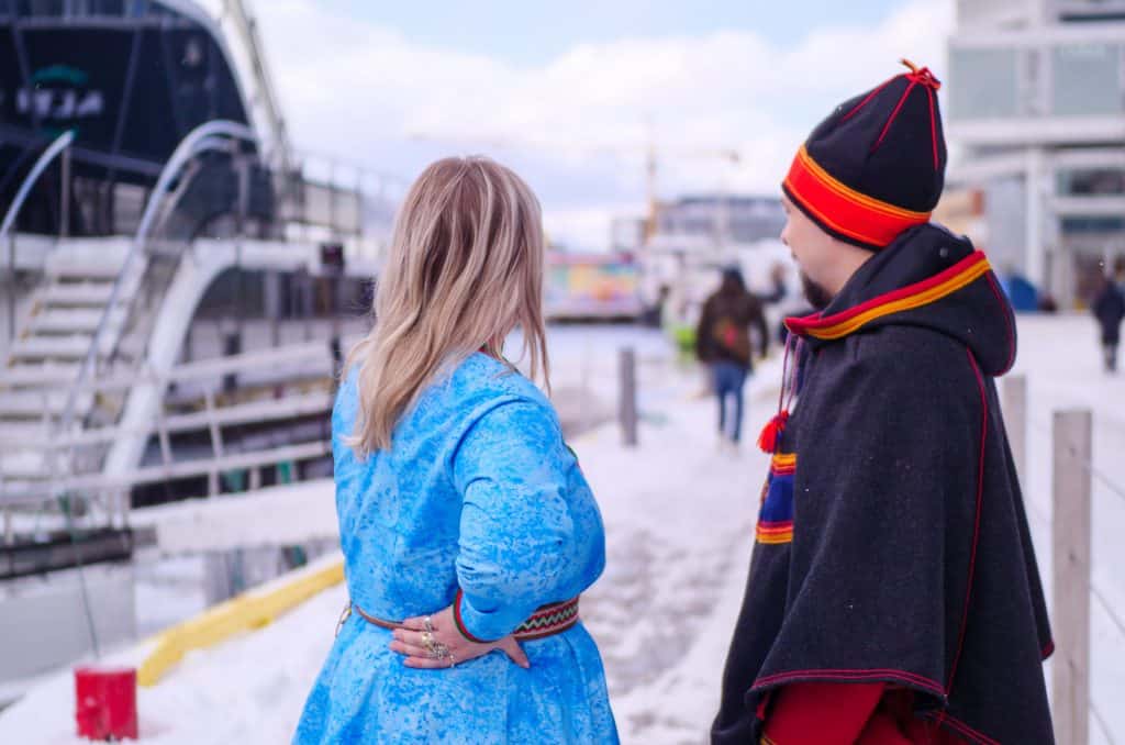 En samisk mann og kvinne i tradisjonelle klesplagg, står ved en havn og ser folk gå langs havnen.