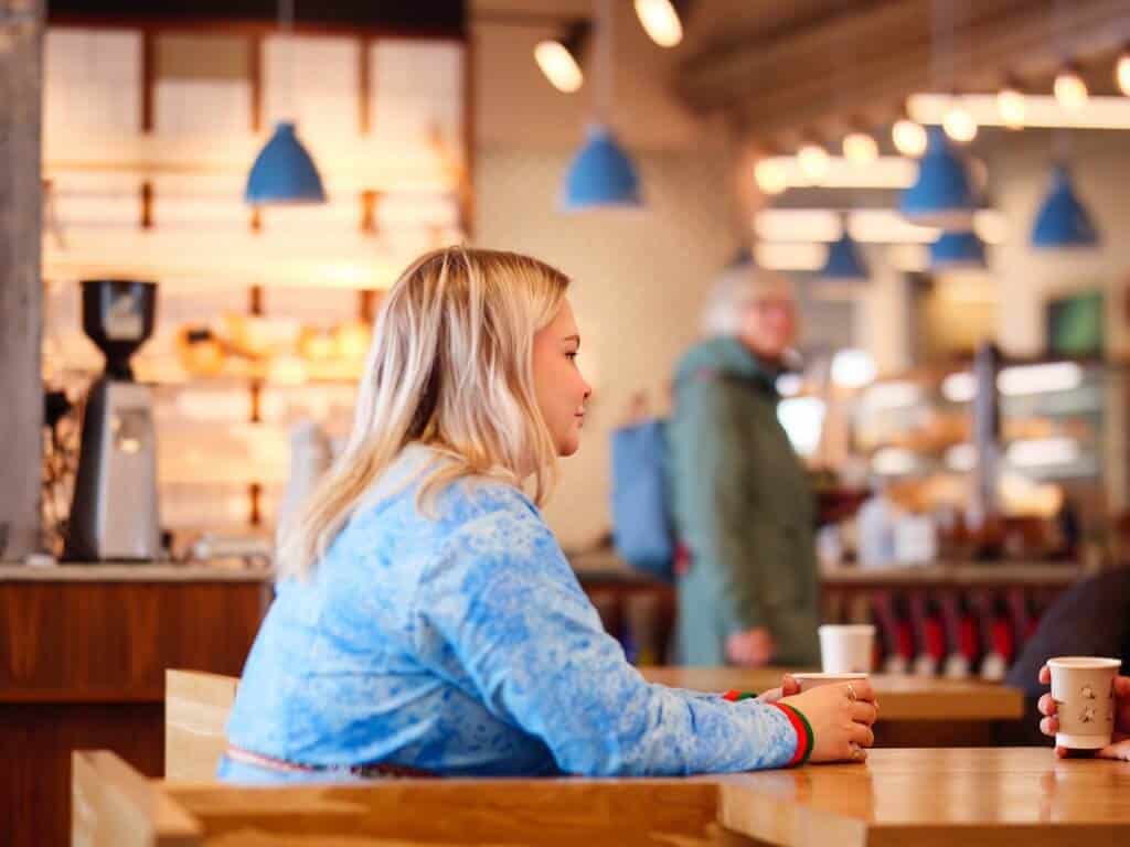 Bildet viser en samisk kvinne sett fra siden, som sitter på en kafé i dialog over en kaffe.