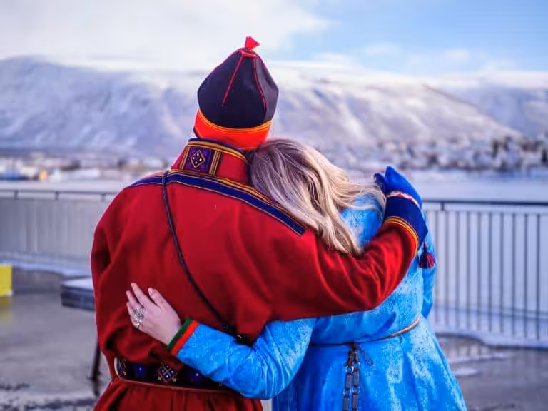 Bilde som viser en samisk mann og kvinne i tradisjonelle kofter. De holder rundt hverandre med Ishavskatedralen og fjellene i Tromsø i bakgrunnen. Kvinnen lener hodet mot mannens skulder.