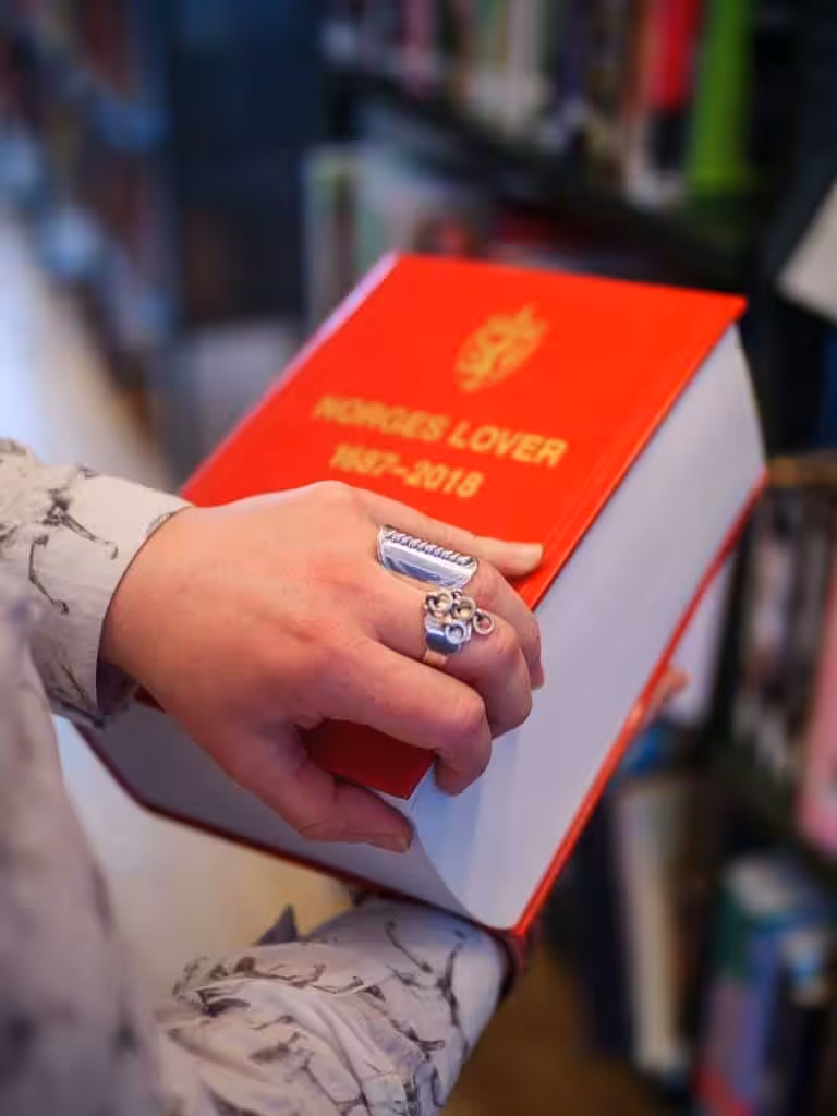 Nærbilde av en samisk kvinnes hender, som er i ferd med å åpne Norges lover. Kvinnen har typiske samiske smykker og armbånd på hendene.