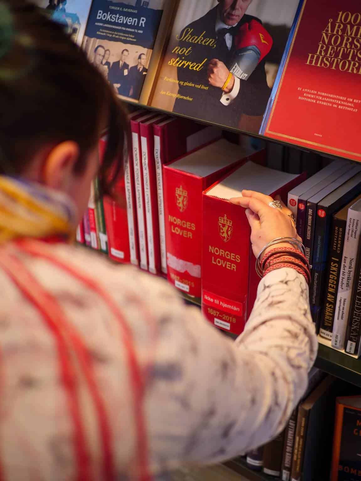 Bilde viser en samisk kvinne med samiske øredobber, ringer og armbånd som tar ut Norges lover fra en bokhylle i biblioteket.