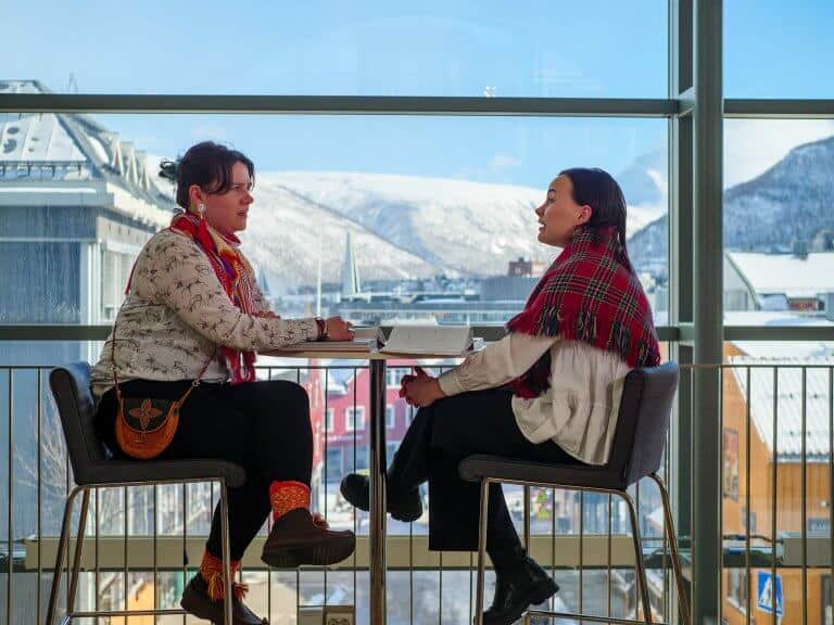 Bildet viser to samiske kvinner i dialog ved et stort vindu. I bakgrunnen ser vi fjellene i Tromsdalen.