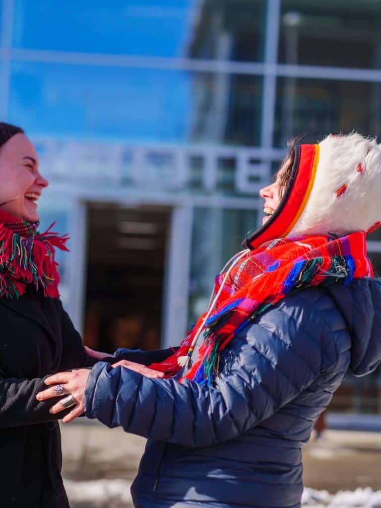 To unge samiske kvinner ler hjertelig, mens de holder hverandre i hendene.